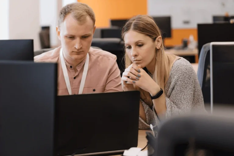 two coworkers focus on a computer screen in a bright office, collaborating at a desk with monitors around them.