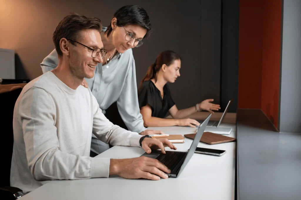colleagues collaborating at a long desk with laptops; man in foreground typing as two coworkers assist nearby.