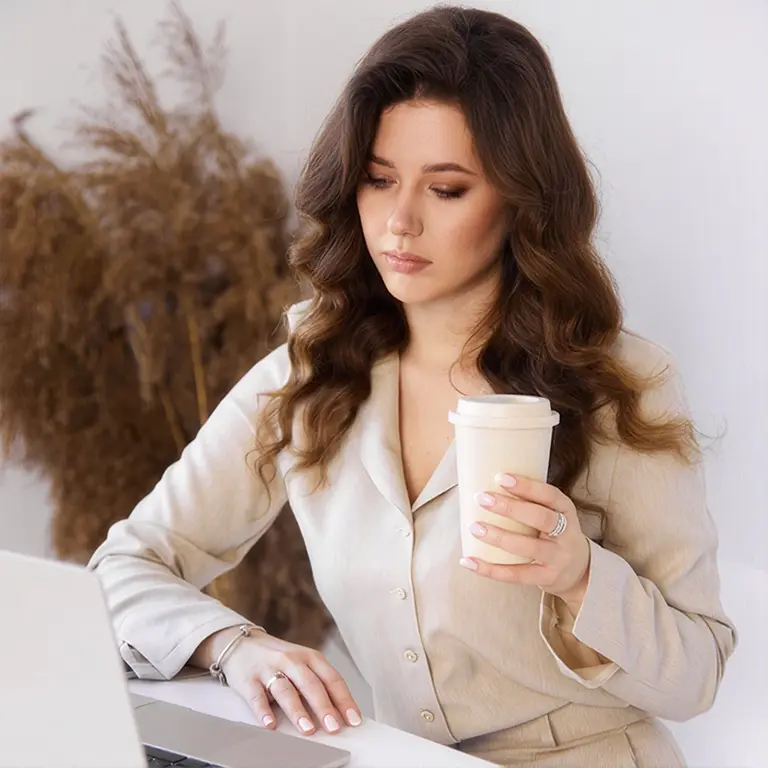 woman with wavy brown hair sits at a desk, holding a to-go coffee cup while looking at a laptop.