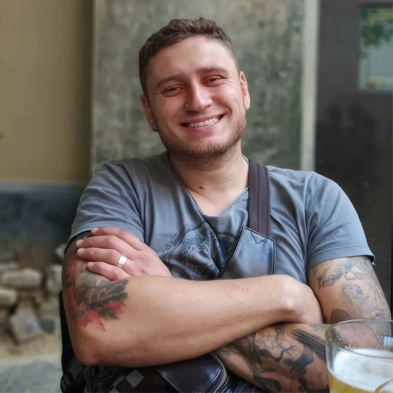 smiling man with tattoos crosses his arms at an outdoor cafe; a beer sits on the table beside him.