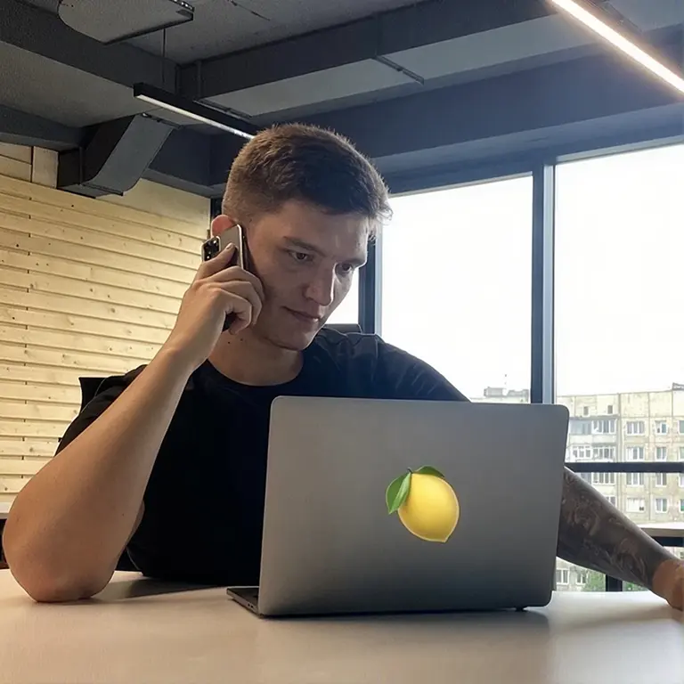 man seated at a desk in an office, talking on a smartphone while looking at a silver laptop with a lemon sticker and a leafy green leaf.",