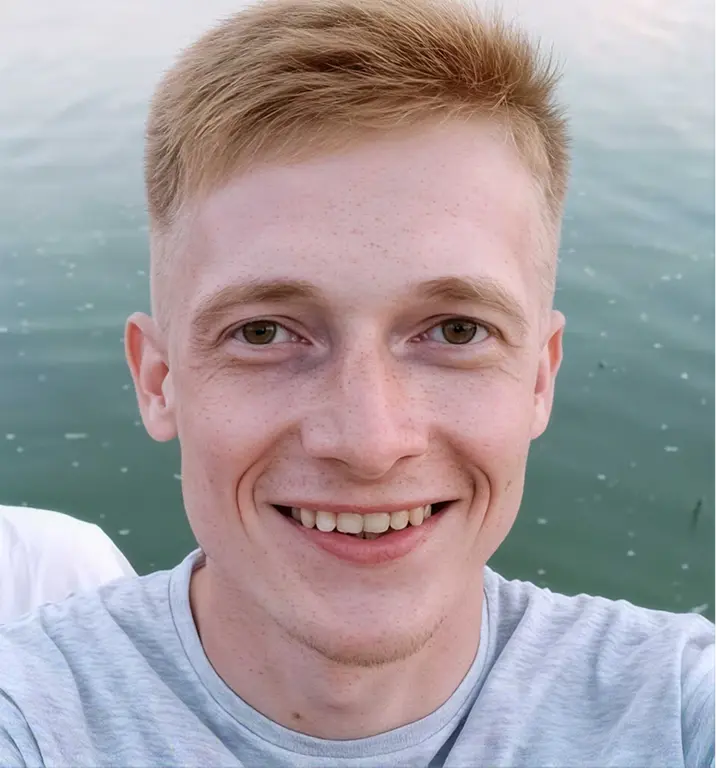 young man with short blond hair and freckles, smiling at the camera outdoors near a body of water.