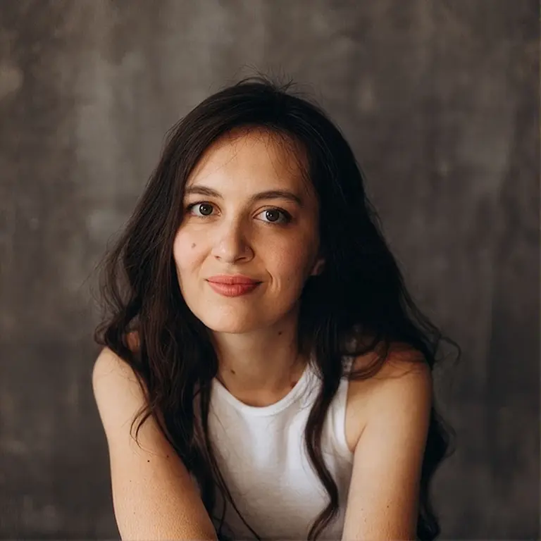 portrait of a young woman with long dark hair, wearing a white sleeveless top, smiling softly against a gray studio backdrop.