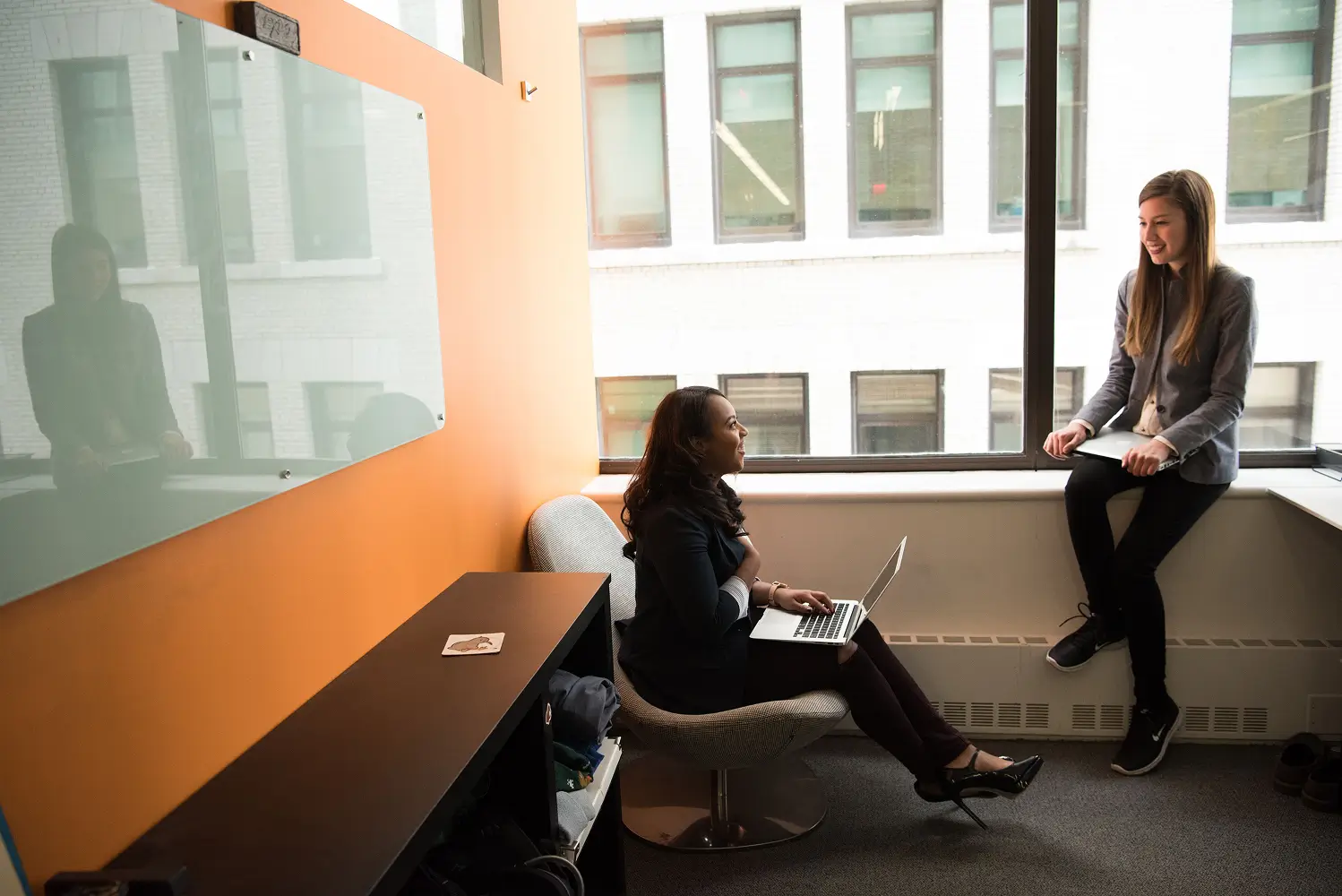 two women in a bright modern office: one seated with a laptop on her lap, the other standing by a window with a notebook, chatting.