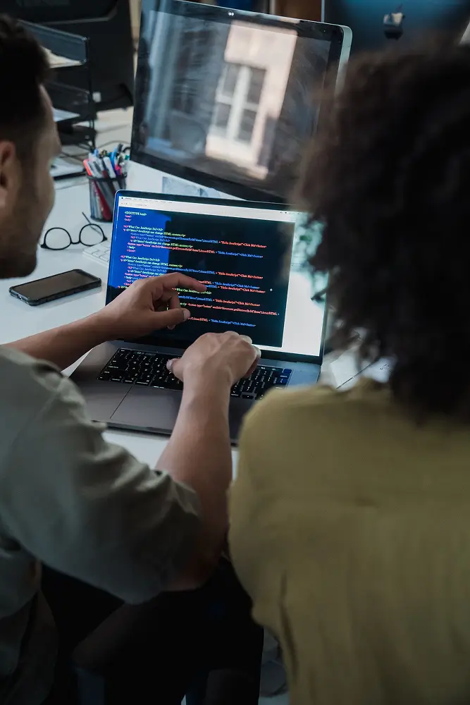 two people pair-programming at a desk, typing code on a laptop screen with colorful text.