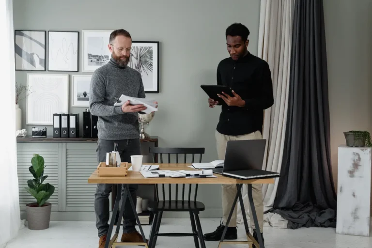 two colleagues review documents at a wooden desk in a modern office, with a laptop and scattered papers nearby in a bright, organized space.