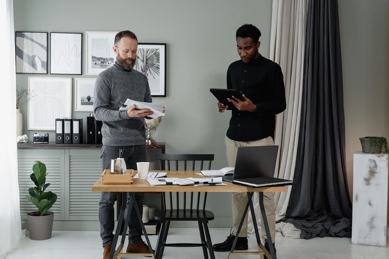 two colleagues review documents at a wooden desk in a modern office, with a laptop and scattered papers nearby in a bright, organized space.