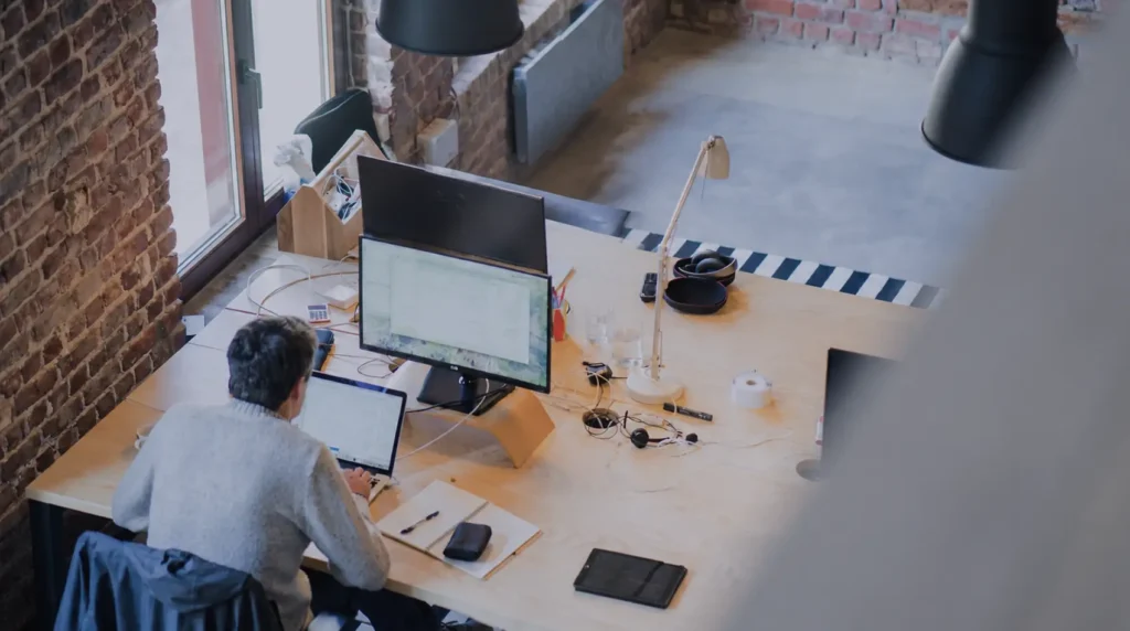 Overhead view of a person working at a wooden desk in a brick-walled loft, typing on a laptop with a large monitor and assorted desk items nearby.