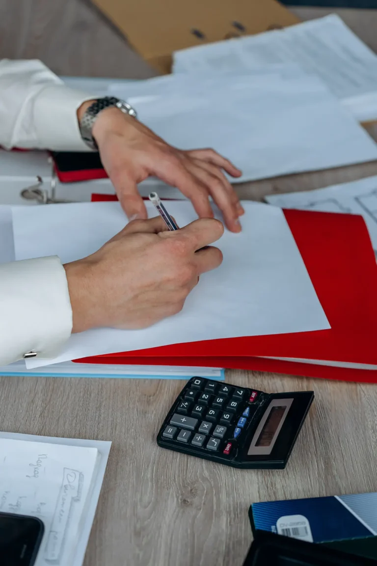 person signing a document with a pen on a desk, surrounded by folders and papers, calculator nearby