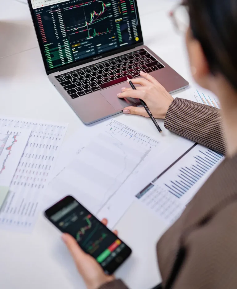 person working at a desk with a laptop showing stock charts, plus printed graphs and a smartphone with a trading app on the desk.